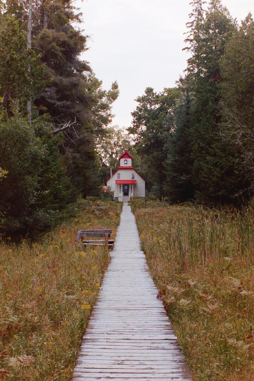 A boardwalk leads through tall grass and trees to a small white building with a red roof.