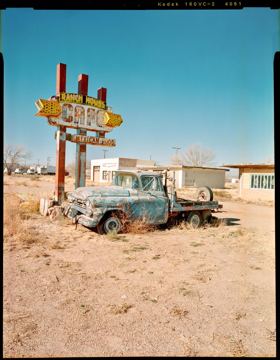 An old, rusty blue truck is parked next to a weathered sign for "Ranch House Cafe Mexican Food" under a clear blue sky.