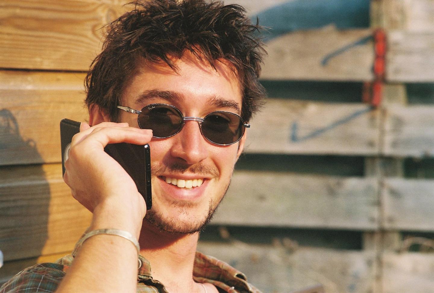 A man wearing sunglasses and a plaid shirt smiles while holding a phone to his ear.