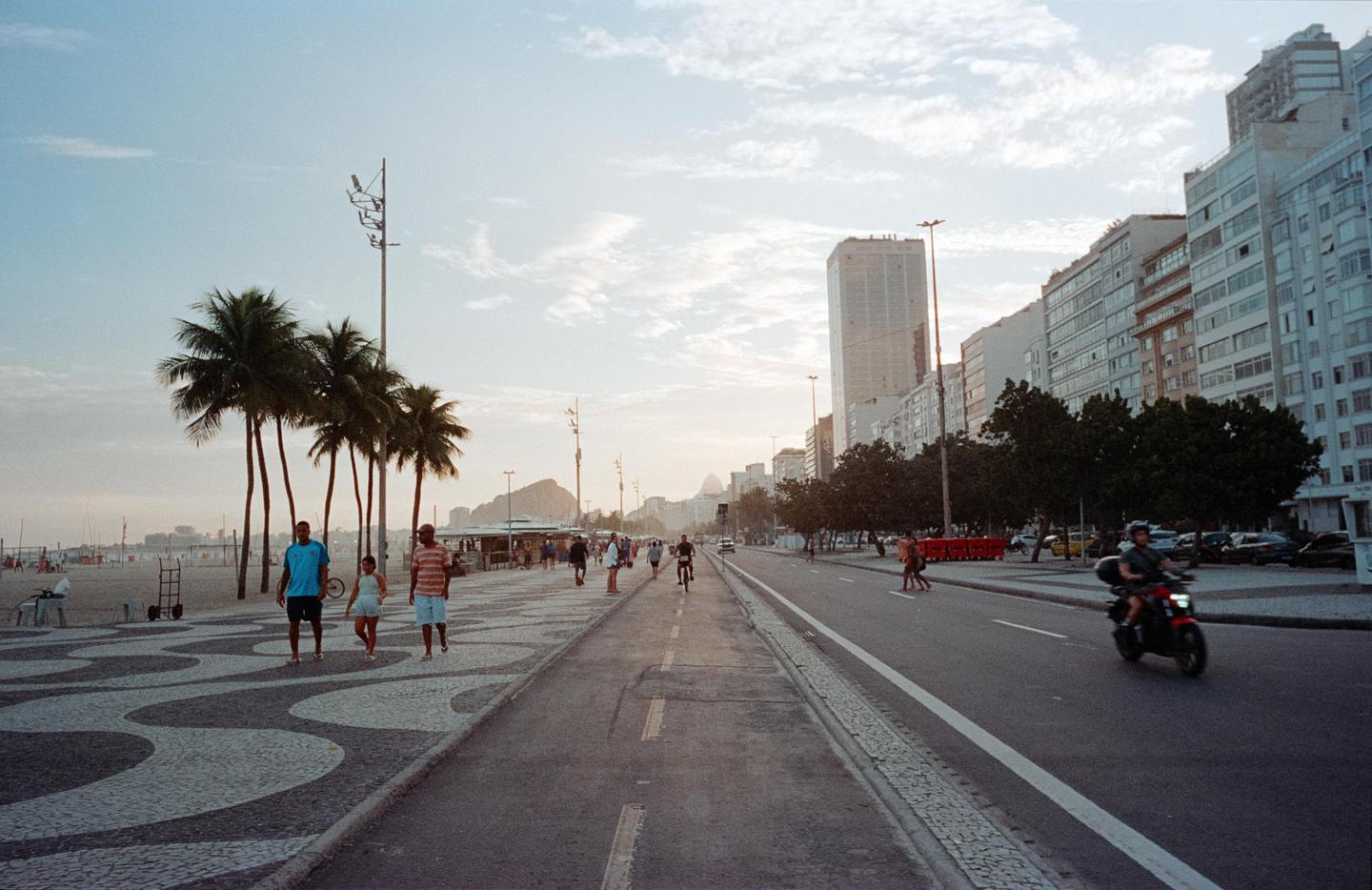 A street with a patterned sidewalk, palm trees, and buildings runs alongside a beach with mountains in the distance.