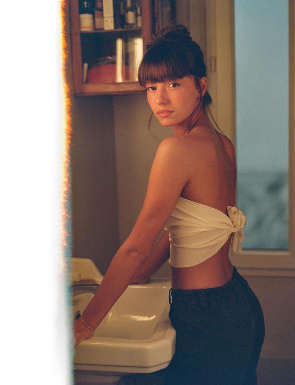 A woman with a bun and bangs looks over her shoulder while leaning on a sink.