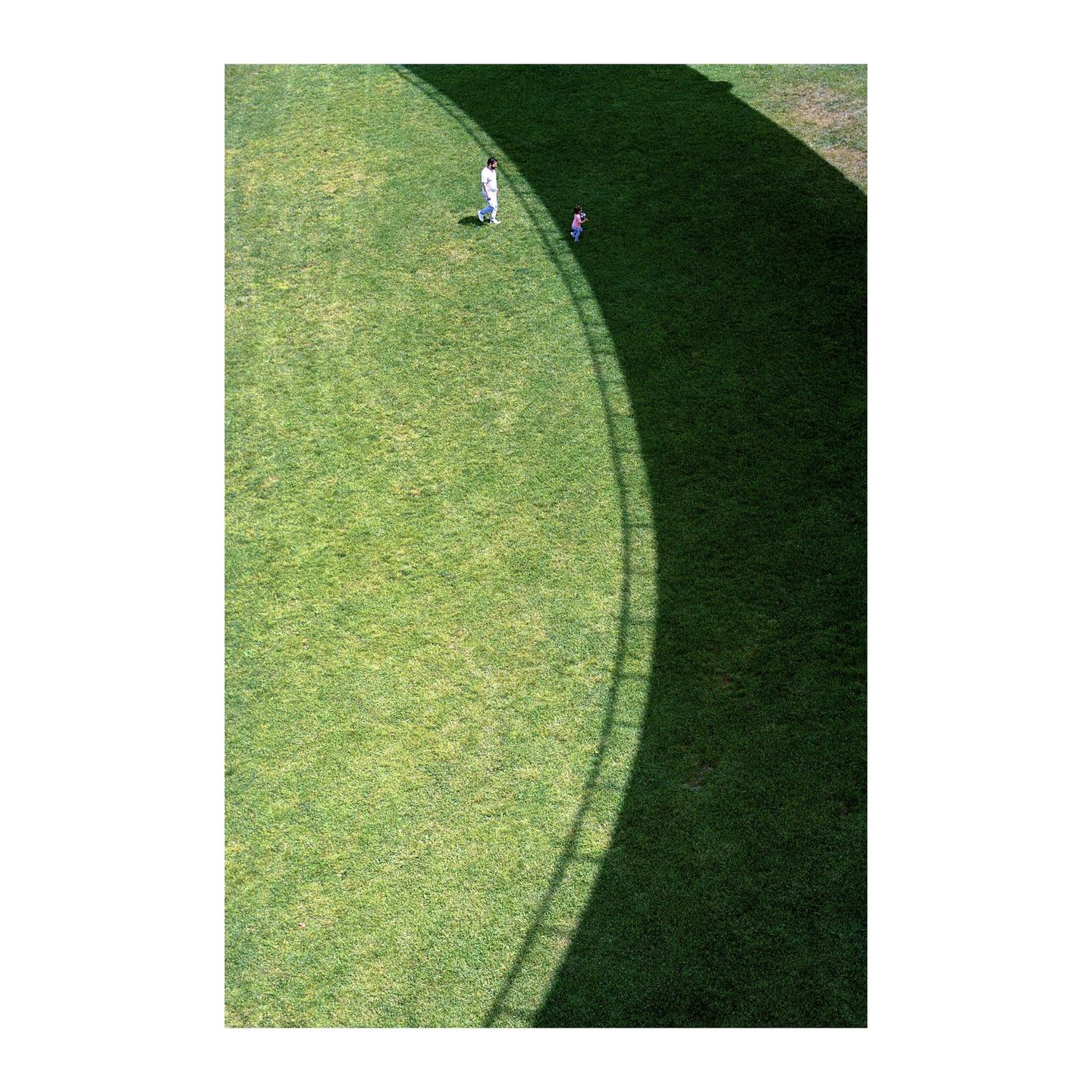 A man and a child walk on a grassy field with a curved shadow.