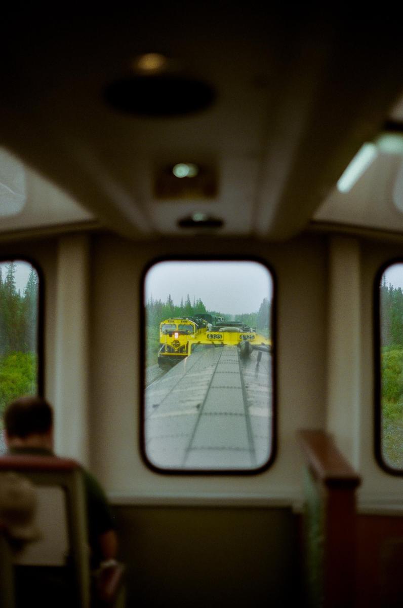 A person sits inside a train car looking out a window at another train on a parallel track.