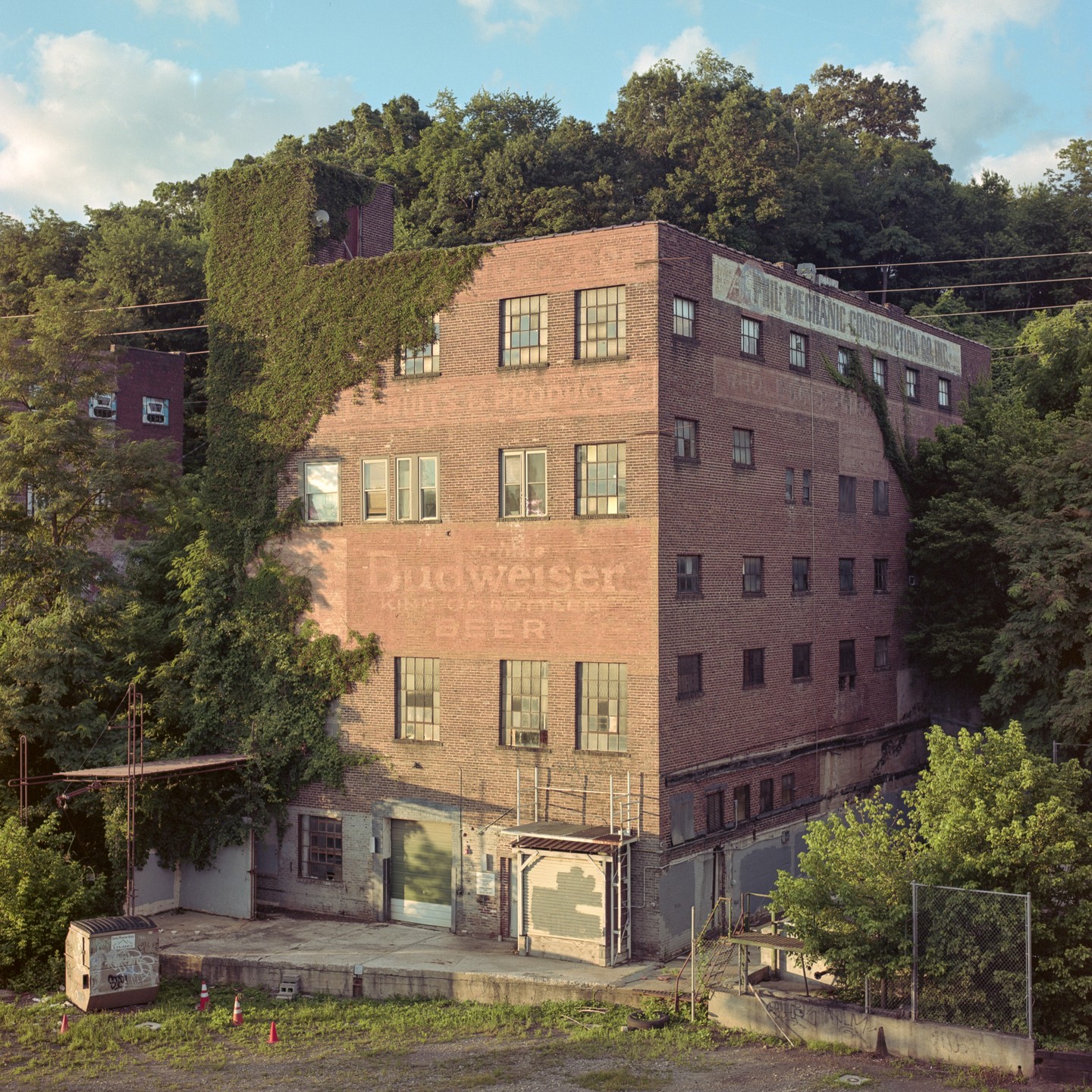 A large brick building with a faded Budweiser advertisement on its side is surrounded by lush green trees.