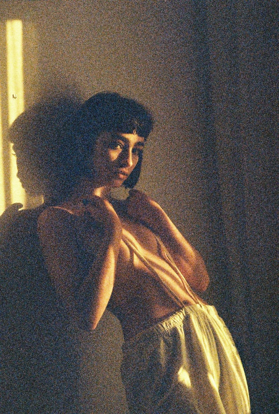 A woman with short hair and bangs is illuminated by a strong light source from the left, casting a shadow on the wall behind her.