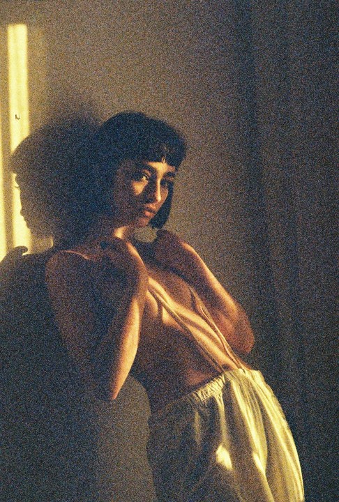 A woman with short hair and bangs is illuminated by a strong light source from the left, casting a shadow on the wall behind her.