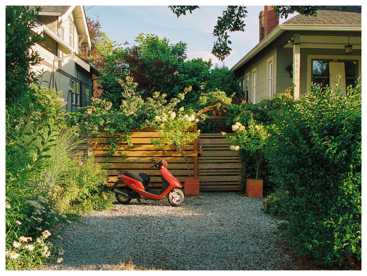 A red scooter is parked on a gravel driveway between two houses and lush green foliage.