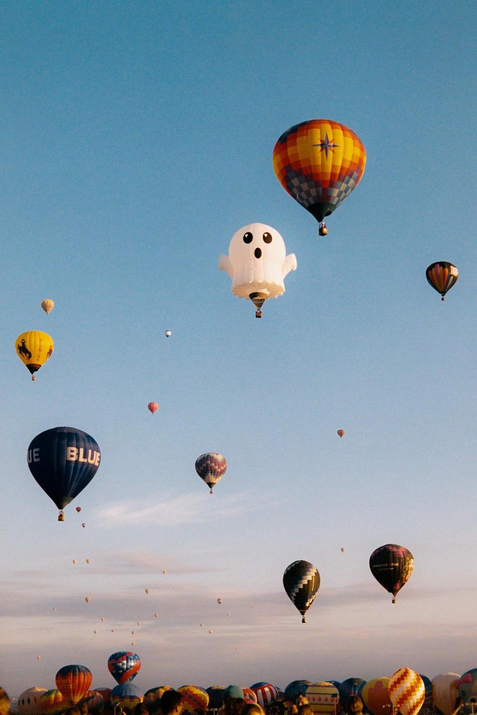 A ghost-shaped hot air balloon floats among many other hot air balloons in a clear blue sky.