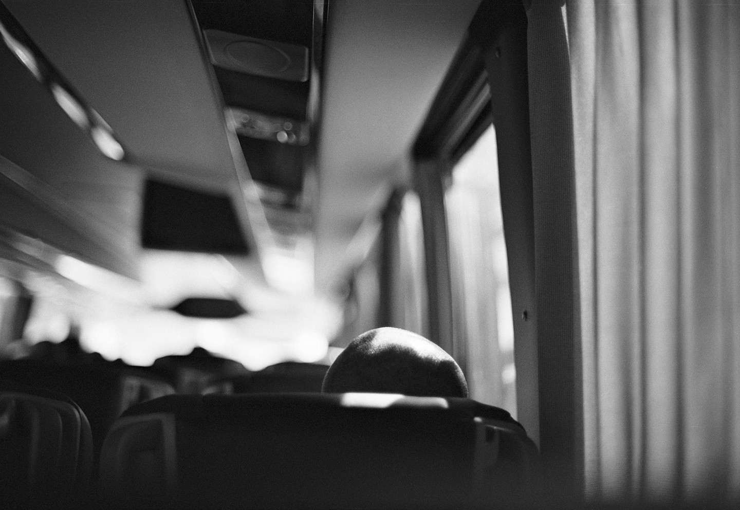 The back of a head is visible above a seat in a vehicle, with a window and curtain to the right.
