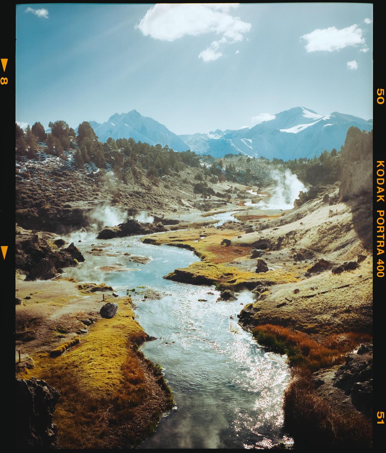 A winding river flows through a valley with steam rising from hot springs, surrounded by mountains and sparse vegetation.