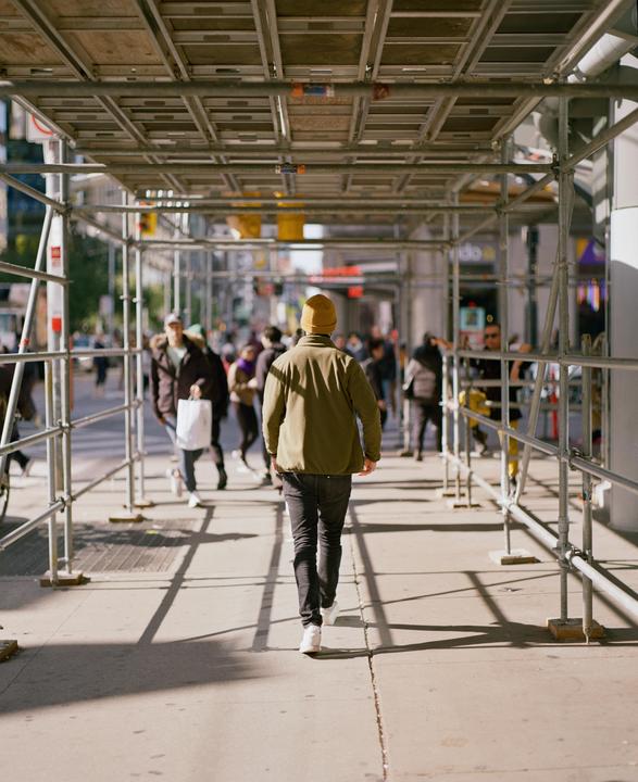 A man in a green jacket and yellow beanie walks away from the viewer under scaffolding on a city street.