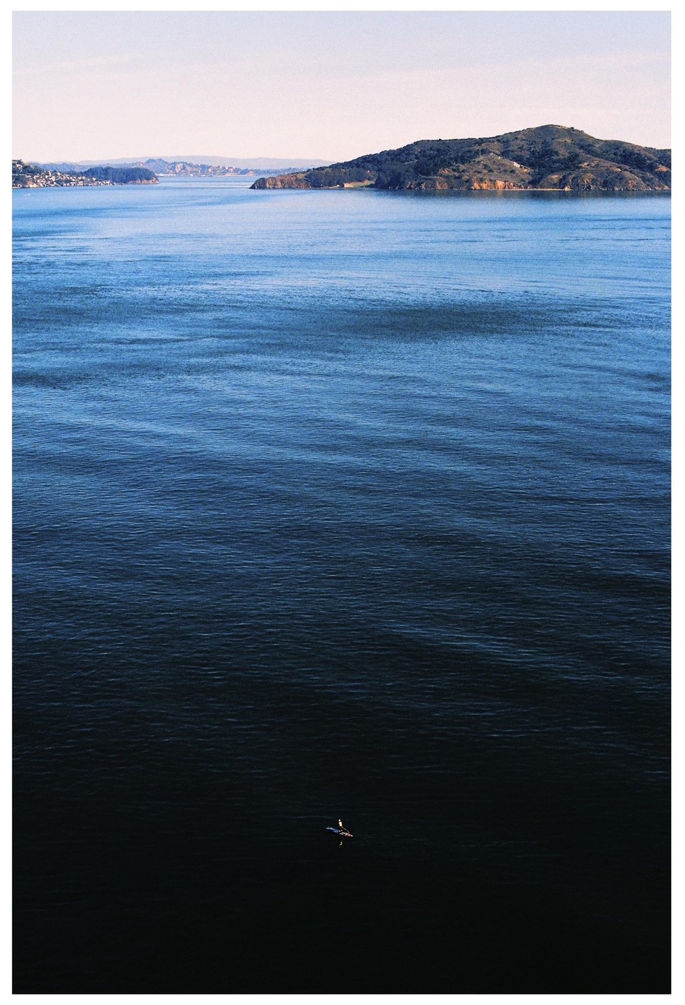 A lone paddler is on the water with a distant island and coastline under a clear sky.