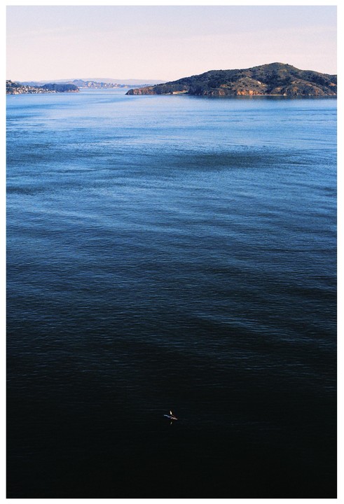 A lone paddler is on the water with a distant island and coastline under a clear sky.