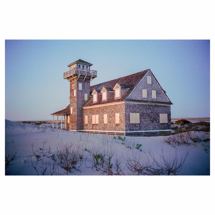 A shingled house with a tower stands on a sandy dune under a clear sky.