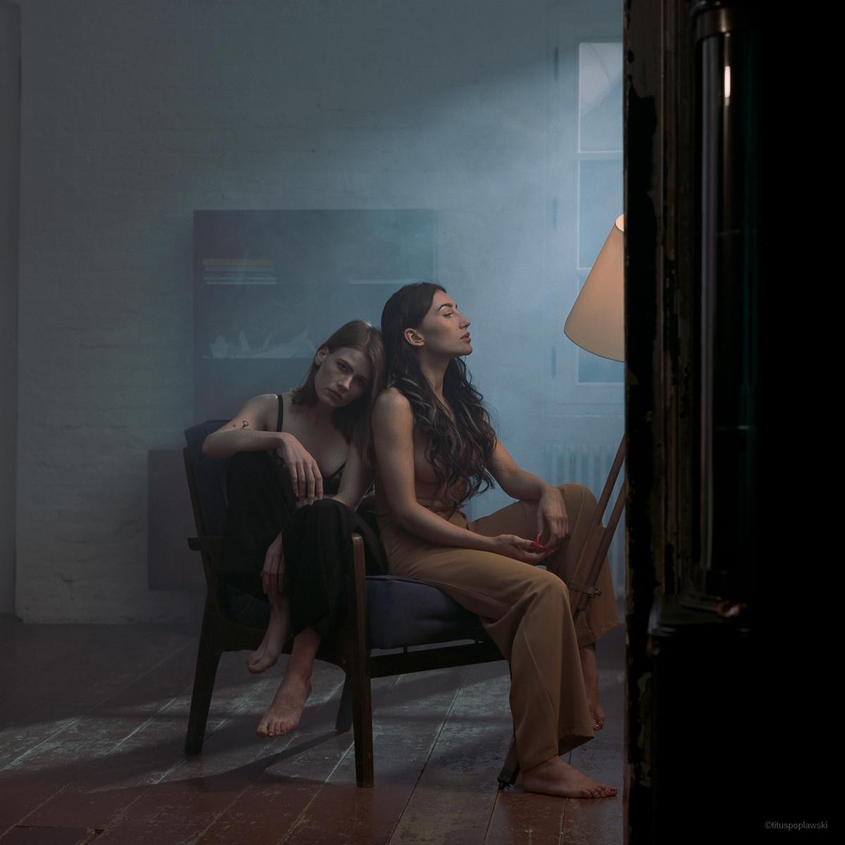 Two women sit in a chair in a dimly lit room with one woman leaning her head on the other's shoulder.