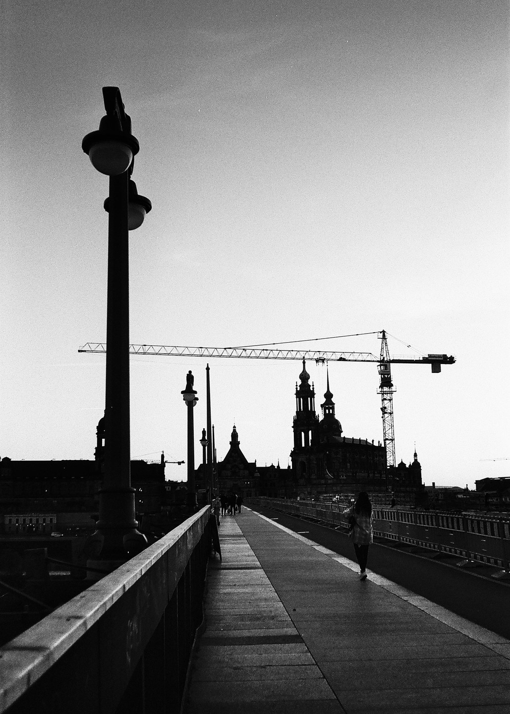 A woman walks along a bridge towards a city skyline with a large crane and historic buildings.