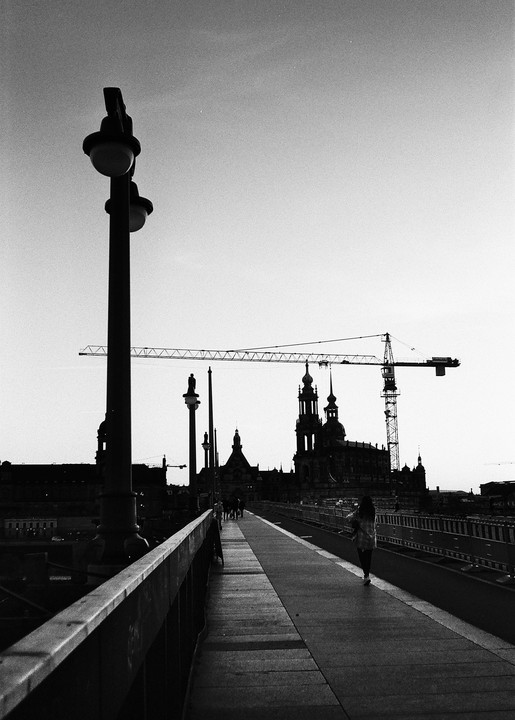 A woman walks along a bridge towards a city skyline with a large crane and historic buildings.