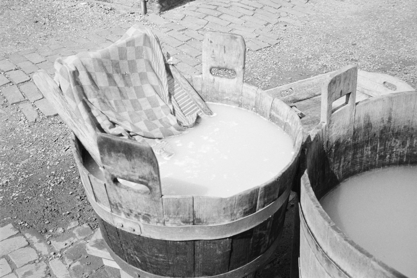 A checkered cloth hangs over the edge of a wooden wash tub filled with water, next to another wash tub.
