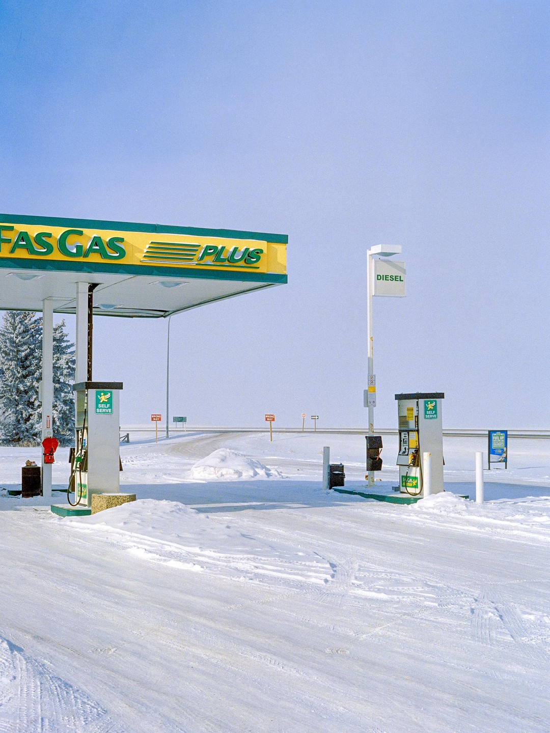 A gas station with a yellow and green sign stands in a snow-covered landscape under a clear sky.