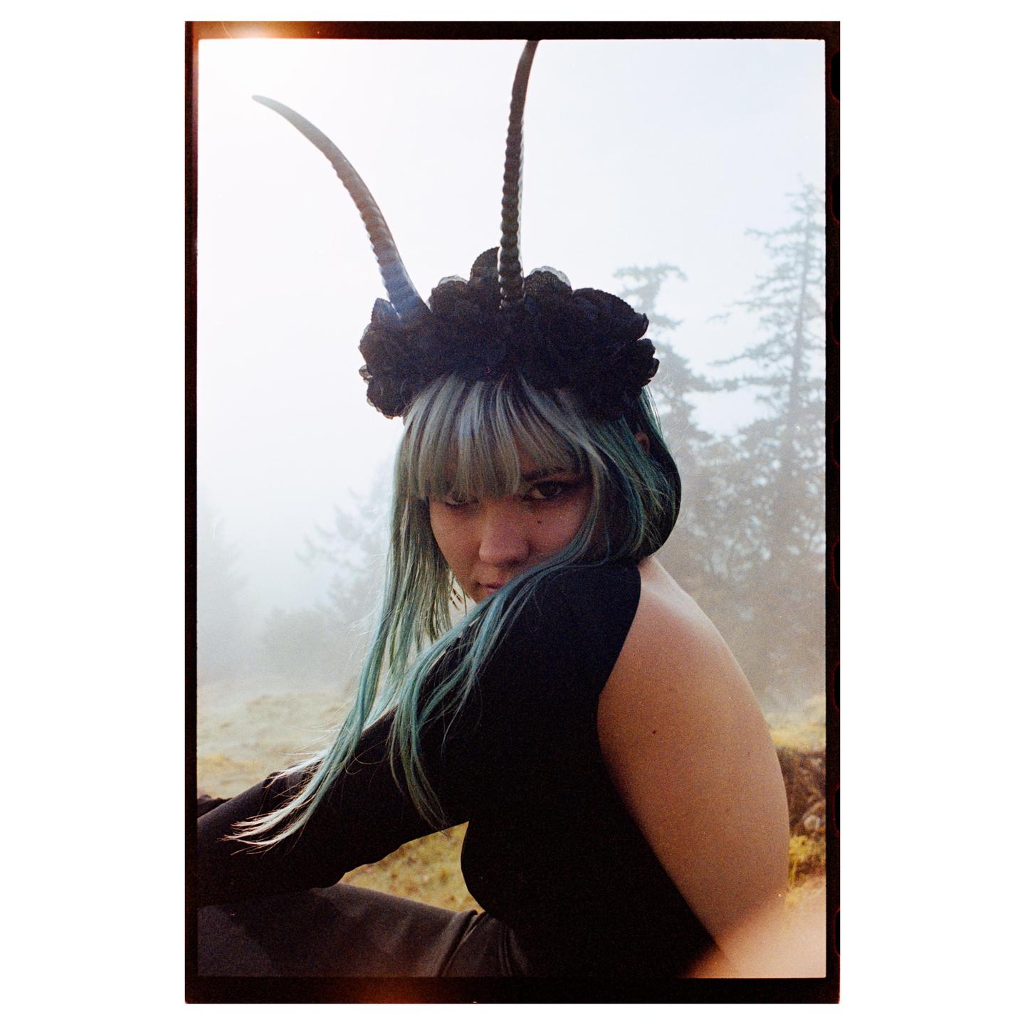 A woman with blue hair and a horned headpiece looks over her shoulder at the viewer in a hazy forest.