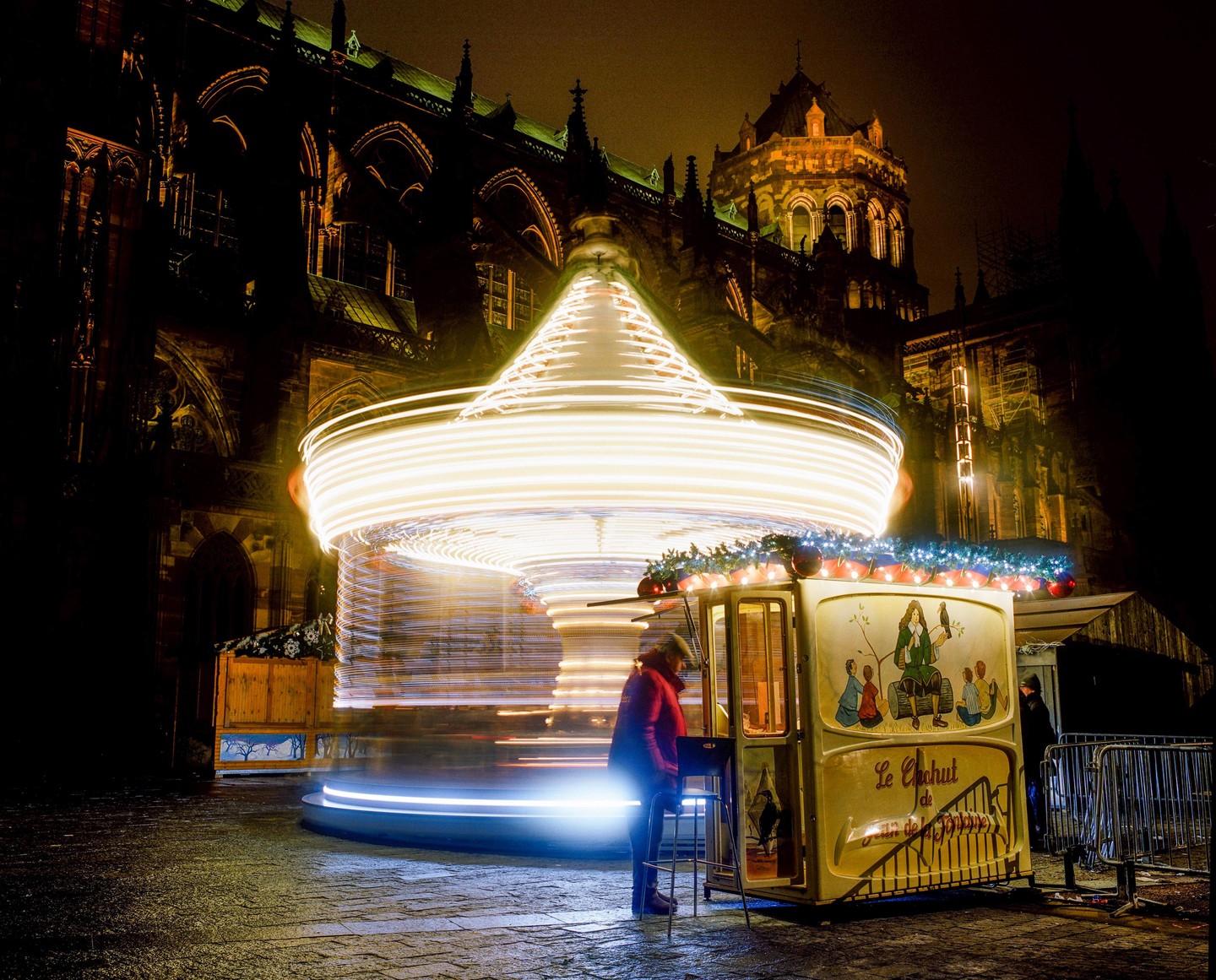 A person stands next to a carousel with light trails in front of a large building at night.