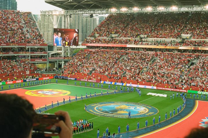 A large crowd of people in a stadium watch a performance on a green field with a large screen displaying a soccer game.