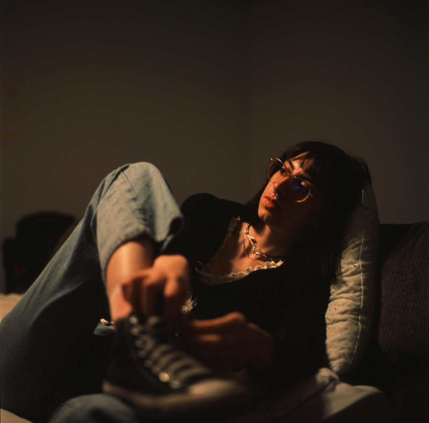 A woman wearing glasses and a necklace is lying on a couch, looking to the side, and holding a shoe.