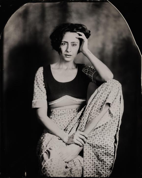 A woman with short, dark hair sits with one leg bent and her hand resting on her head.