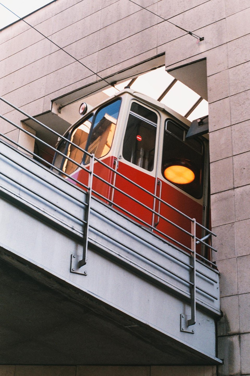 A red and white funicular car is visible through an opening in a modern building with a metal railing in the foreground.
