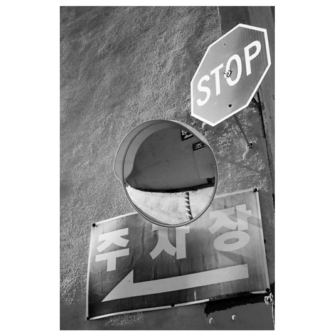 A convex mirror reflects a striped pole below a stop sign and a sign with an arrow and Korean characters.