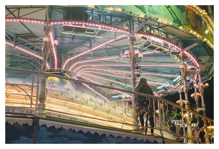 A woman stands on a platform overlooking a brightly lit carousel at night.