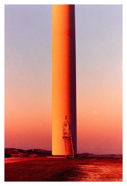 A wind turbine stands in a field at sunset, with a ladder leading to a door on its base.