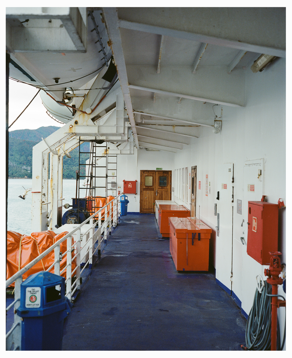 A ferry deck with orange life rafts and red storage boxes is visible next to a body of water with mountains in the distance.