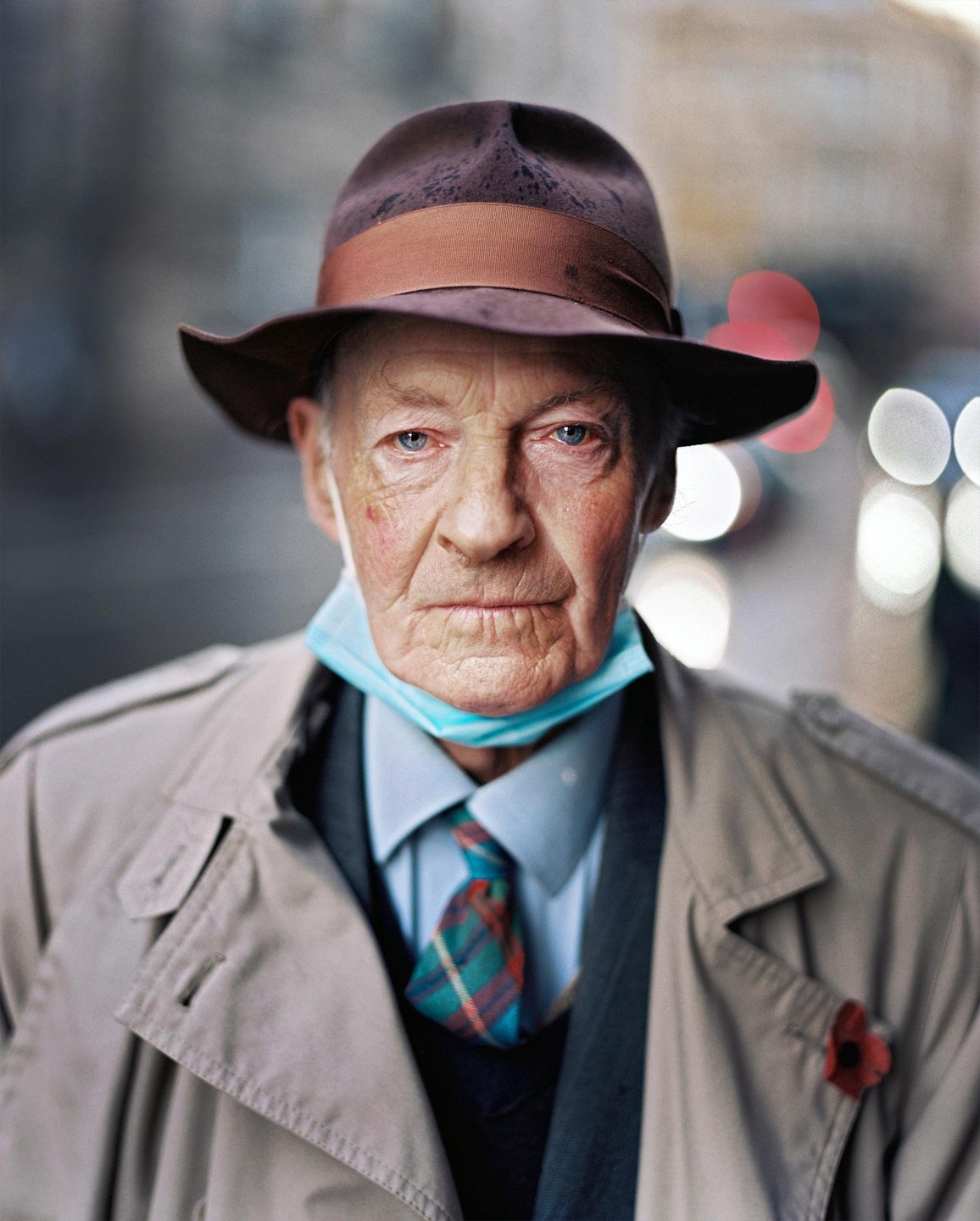 A man wearing a hat, trench coat, and face mask looks directly at the viewer.