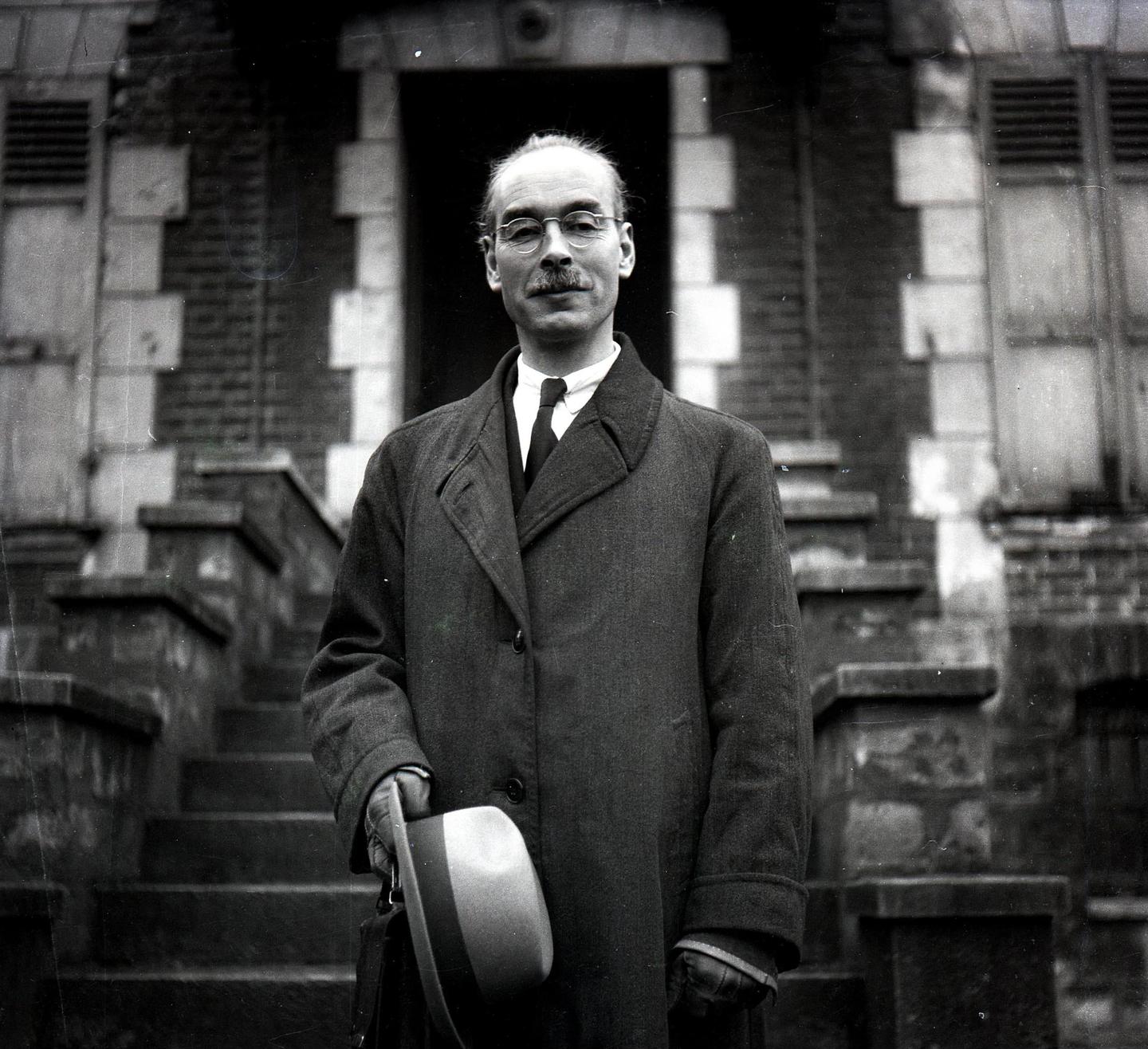 A man in a coat and tie stands on a stone staircase, holding a hat.