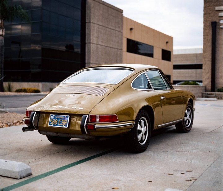 A gold Porsche is parked on a concrete surface in front of a modern building.