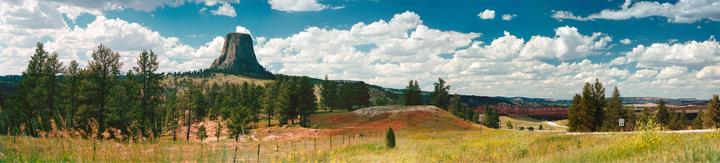 A panoramic landscape shows Devils Tower rising above a forest and fields under a blue sky with white clouds.