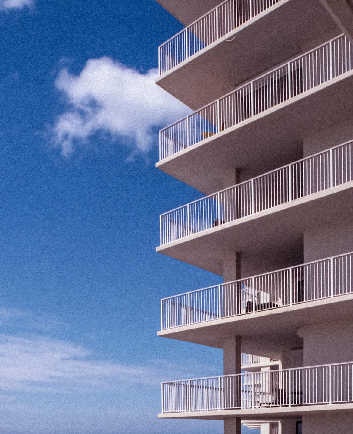 A tall building with many balconies is shown against a bright blue sky with a single cloud.
