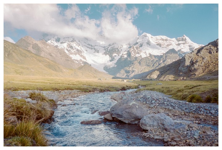 A river flows through a valley with snow-capped mountains in the background under a cloudy sky.