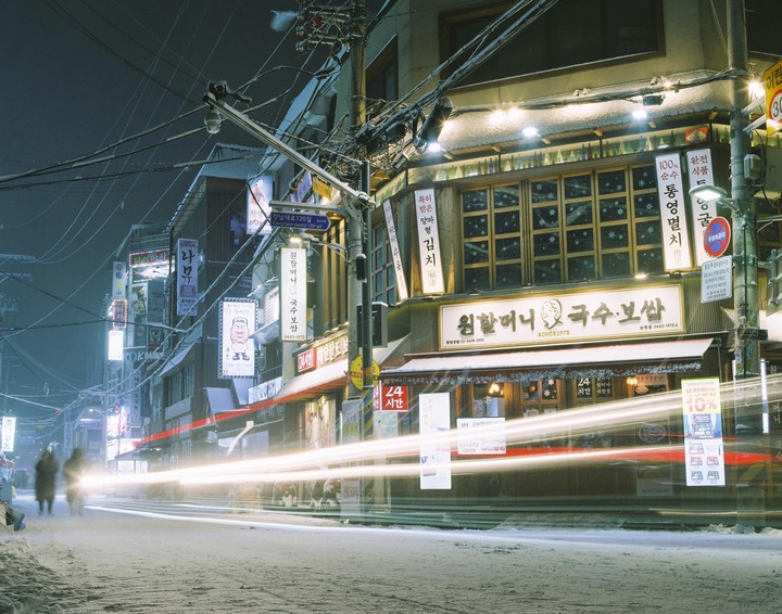 A snow-covered street at night is lined with buildings displaying illuminated signs, with light trails from passing vehicles.