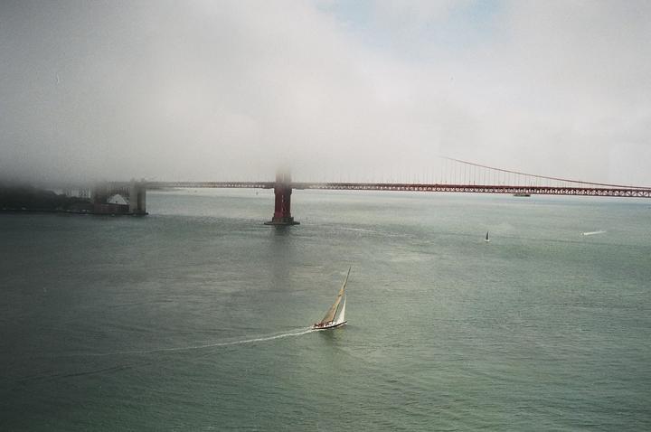 A sailboat moves across the water with the Golden Gate Bridge partially obscured by fog in the background.