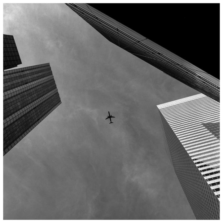 An airplane flies high above, framed by three tall buildings.
