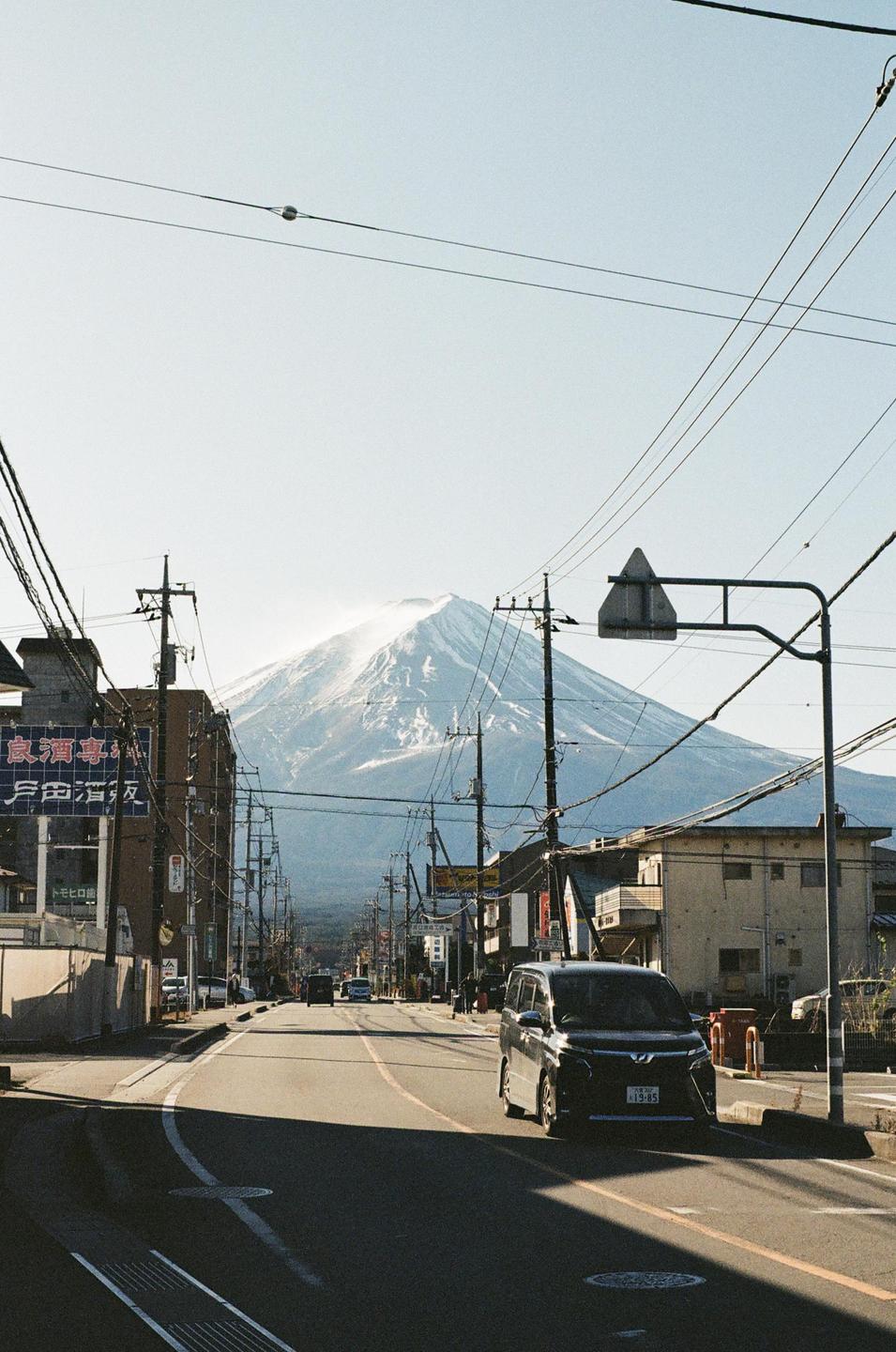 A snow-capped mountain rises above a street with buildings and power lines.