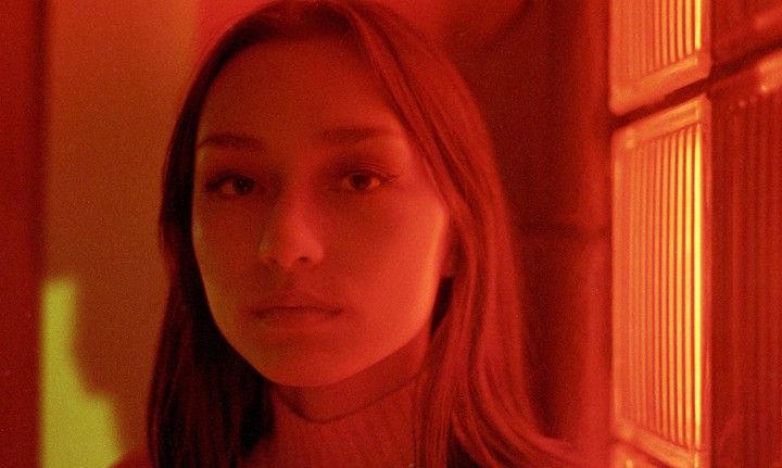 A woman with long hair looks directly at the viewer, illuminated by red and yellow light.