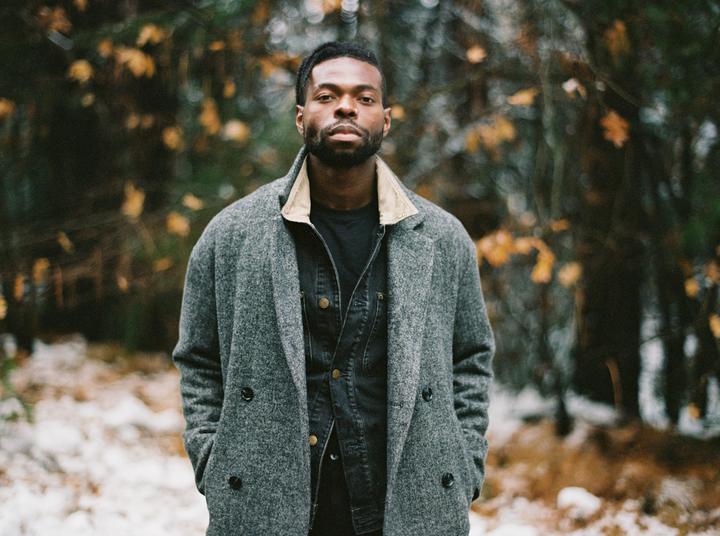 A man wearing a gray coat and a denim jacket stands in a forest with patches of snow on the ground.