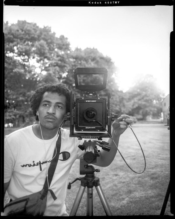 A man with curly hair and a white t-shirt stands next to a large format camera on a tripod in a grassy area with trees in the background.