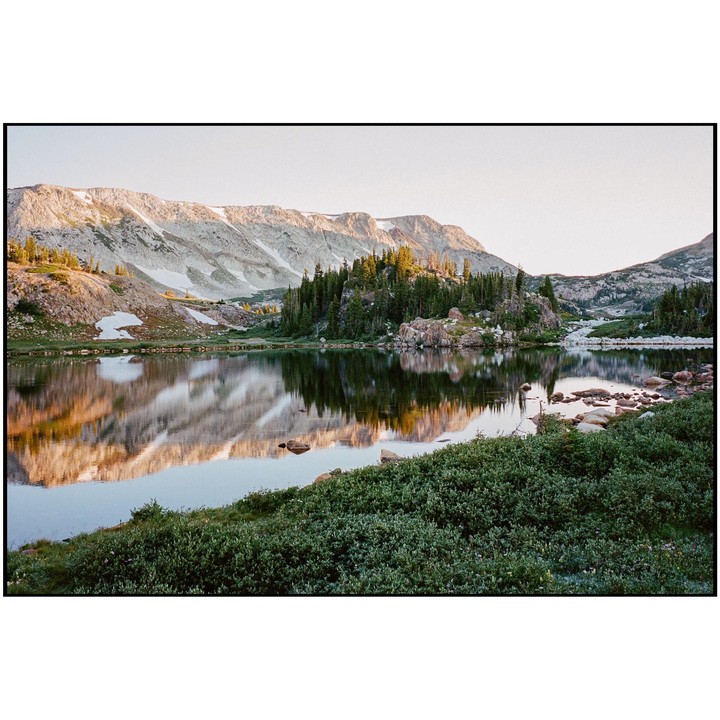 A mountain range with snow-capped peaks is reflected in a calm lake surrounded by evergreen trees and green bushes.