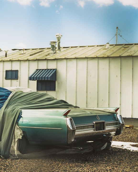A teal vintage car with a partial cover is parked on a gravel surface in front of a white building.