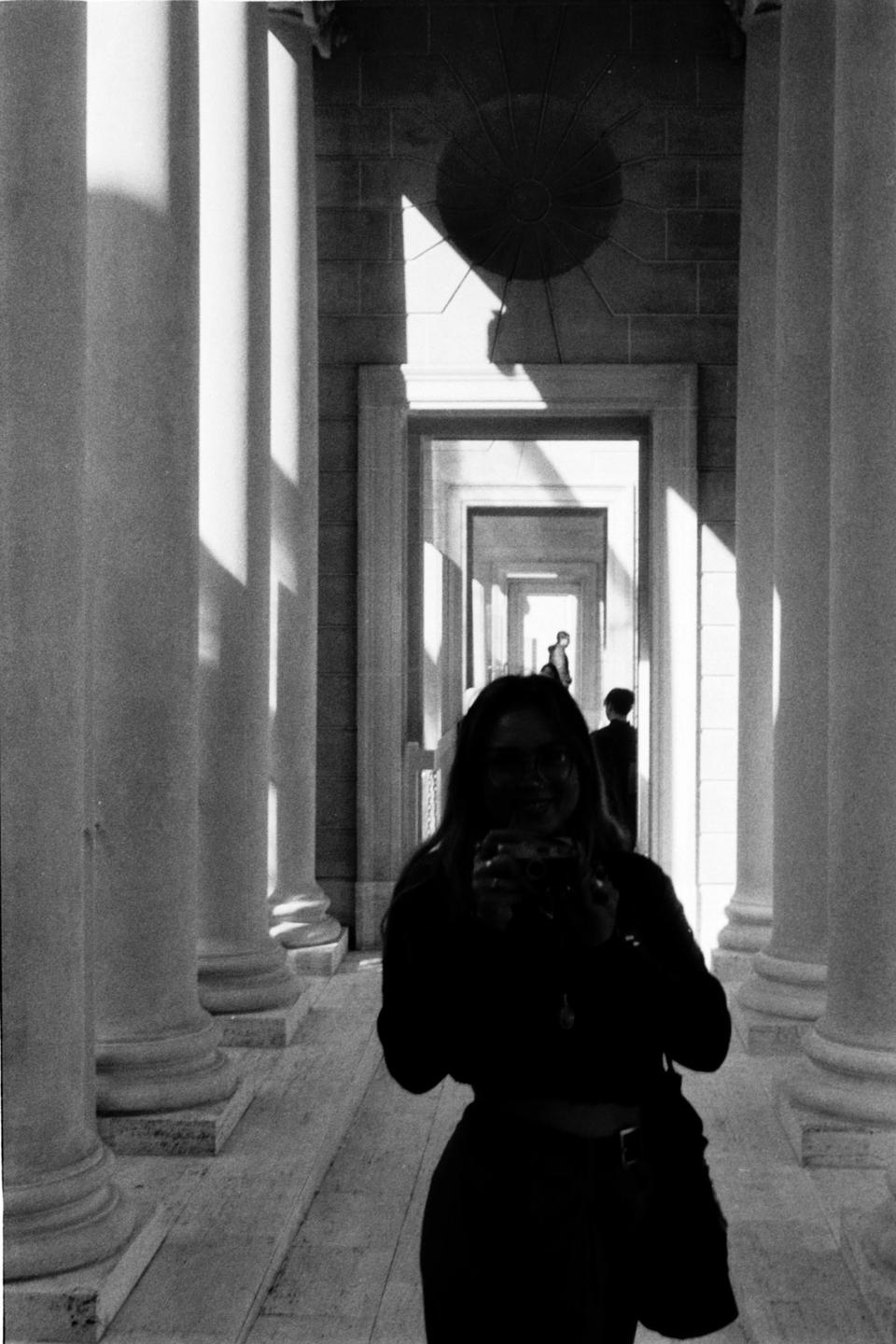 A woman takes a self-portrait in a long hallway with columns and doorways, with another person visible in the distance.
