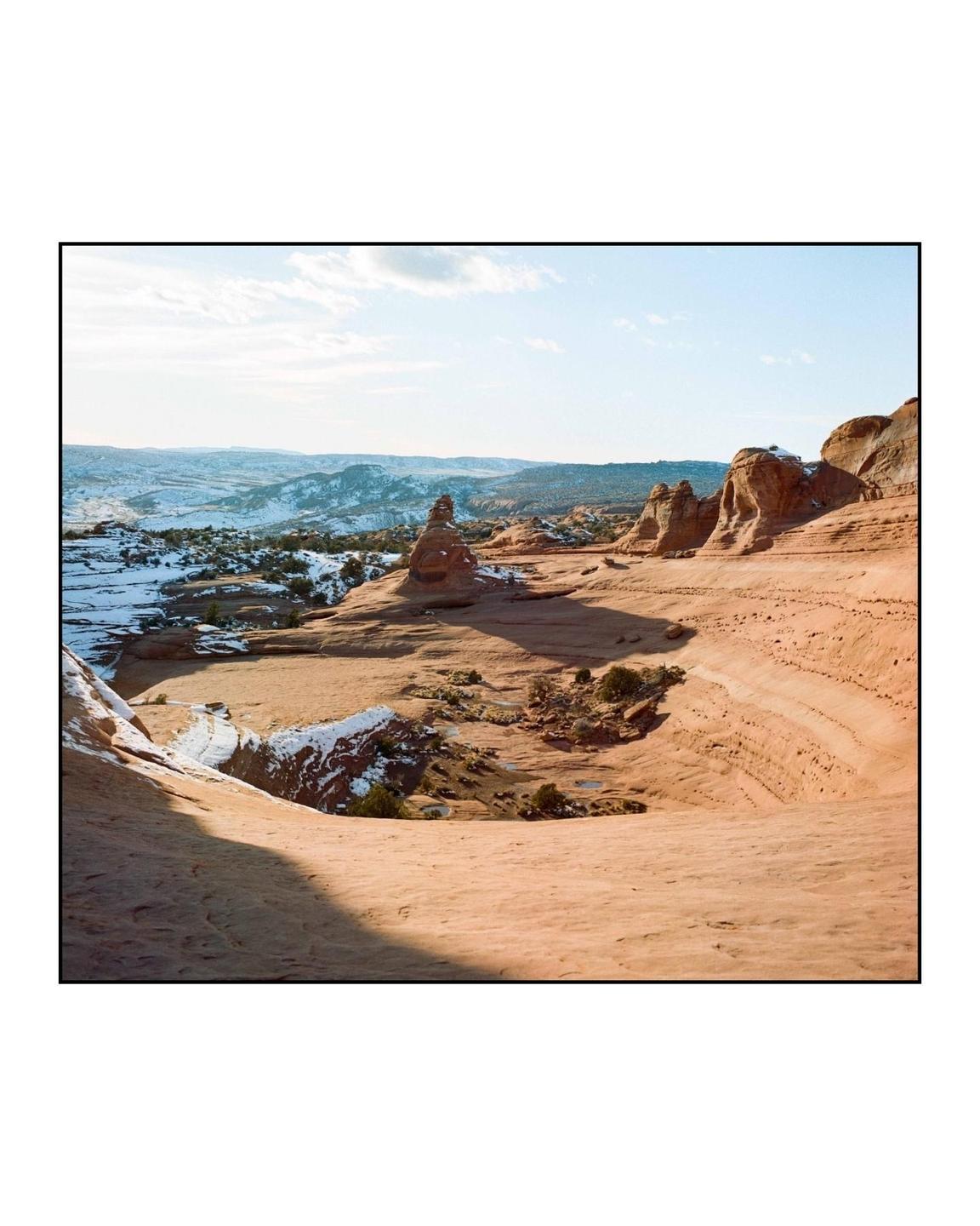 A vast desert landscape with red rock formations and patches of snow is visible under a clear sky.